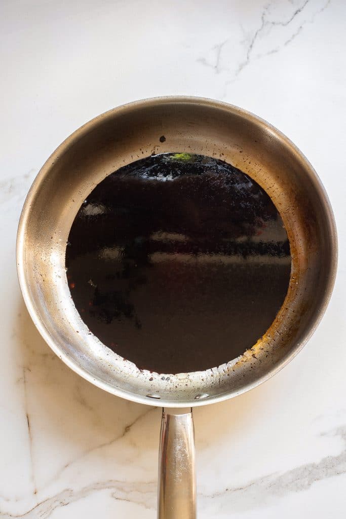 A stainless steel frying pan with a dark, glossy liquid covering the bottom, possibly a sauce or oil, is placed on a white marble surface. The pan has a slight sheen, and the liquid appears to be thick and reduced.