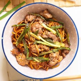 A bowl filled with stir-fried noodles topped with cooked ground meat and green vegetables. The dish is set on a light-colored table with bamboo and green garnish alongside. The noodles appear glossy, suggesting a savory sauce.