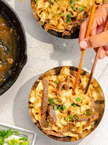 Two bowls of stir-fried noodles with beef and scallions are placed on a light surface. A hand with chopsticks is picking up a portion from one bowl. A cast iron skillet with sauce and a small dish of chopped scallions are nearby.