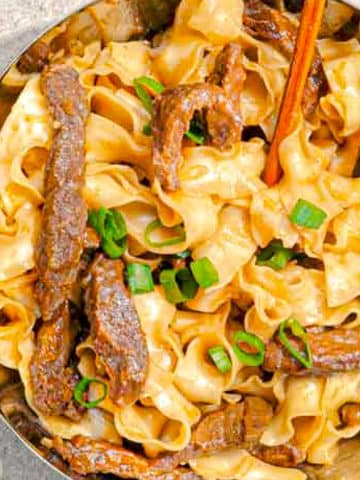 A metallic bowl filled with wide, ribbon-like noodles topped with sliced beef strips and garnished with chopped green onions. A pair of wooden chopsticks is placed in the bowl, and a portion of another dish is partially visible on the side.
