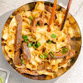 A bowl of wide, flat noodles mixed with seasoned beef strips, garnished with chopped green onions. Two wooden chopsticks are resting on the edge of the bowl. The dish appears flavorful and well-presented.