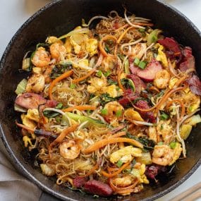 A skillet filled with a stir-fry dish consisting of shrimp, sliced barbeque pork, vegetables including carrots, green onions, and bean sprouts, and vermicelli noodles. The dish is seasoned and cooked with visible egg pieces mixed throughout.