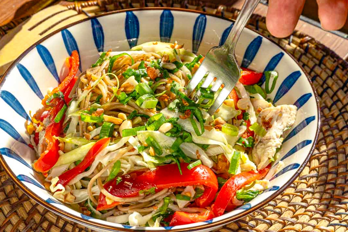 A colorful bowl of noodle salad with sliced red bell peppers, shredded cabbage, green onions, and peanuts. The salad is garnished with herbs and is being mixed with a fork held by a hand. The bowl features a blue and white pattern and rests on a woven mat.