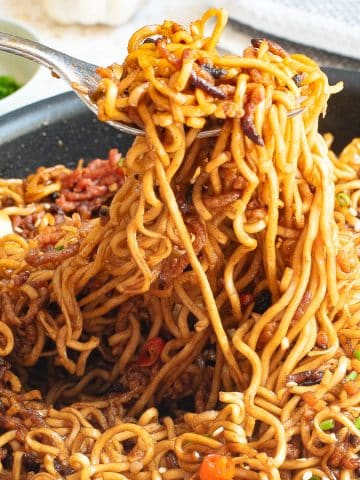 A close-up of a pan filled with stir-fried noodles mixed with vegetables and sauce. A fork is lifting a portion of the noodles, showing their texture and the various ingredients, including chunks of vegetables and seasonings.