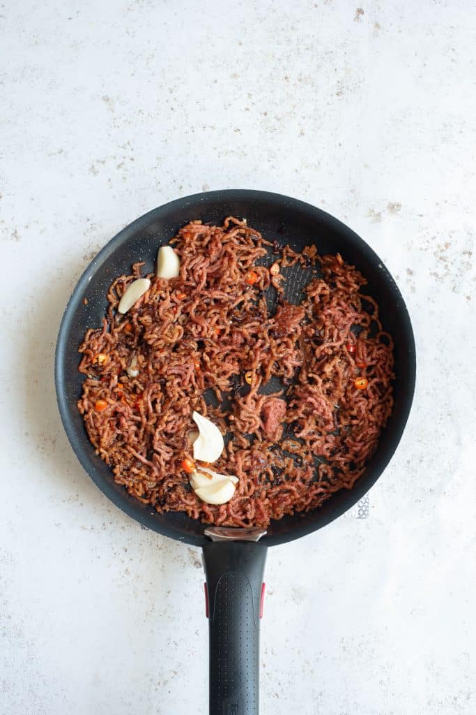 A black frying pan on a light-colored surface contains cooked ground beef mixed with small chunks of garlic and finely chopped onions and vegetables. The beef appears browned and well-cooked.