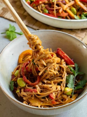 A close-up of a bowl of peanut noodle salad featuring colorful ingredients such as red bell peppers, edamame, carrot shreds, and cilantro. Chopsticks are lifting a twirl of noodles, and another bowl of salad sits in the background.