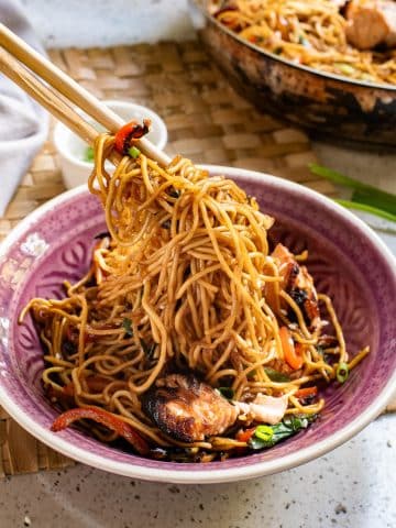 A bowl of stir-fried noodles with vegetables and pieces of meat is being lifted by a pair of chopsticks. The bowl is purple on the inside with a light pink rim. Various veggies like bell pepper and greens are visible among the noodles.
