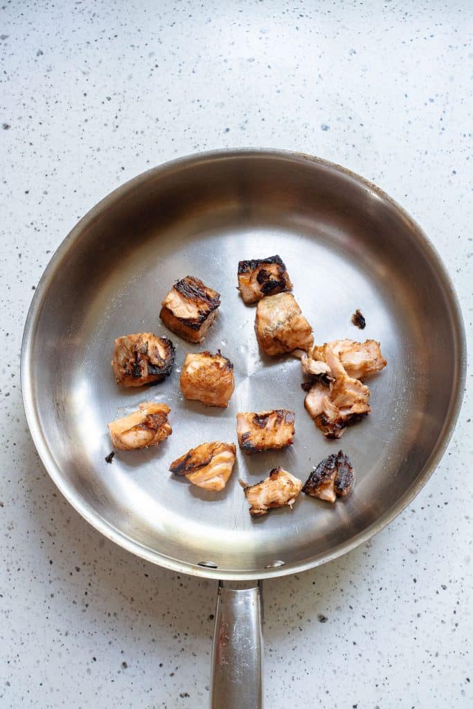 A stainless steel frying pan containing several small, browned pieces of cooked meat scattered inside. The pan rests on a light-colored, speckled countertop. Some pieces have slightly charred edges, indicating they have been thoroughly cooked.