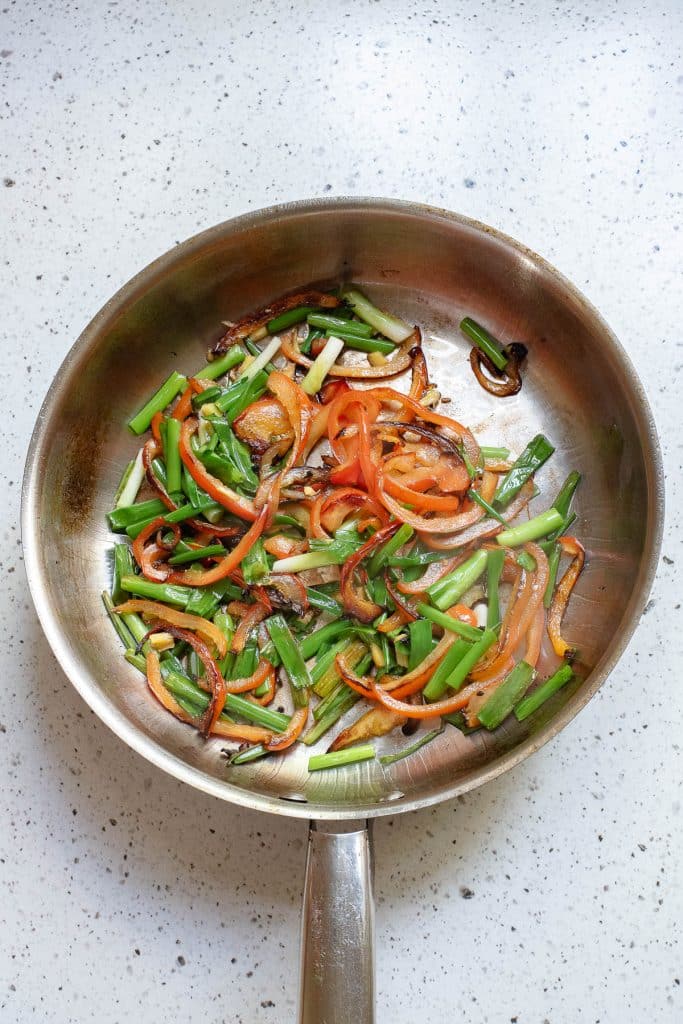 A skillet containing sautéed green onions, bell pepper slices, and onions, resting on a speckled countertop. The vegetables are lightly browned and caramelized.