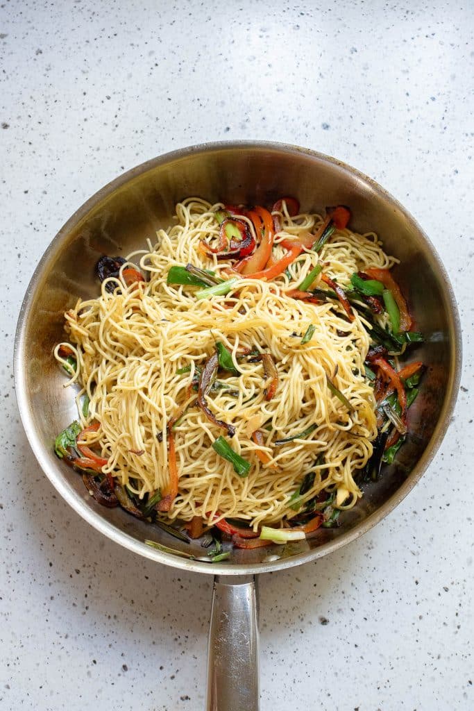 A stainless steel frying pan on a speckled countertop contains a stir-fry of thin noodles mixed with sliced vegetables, including red bell peppers, green beans, and possibly mushrooms. The dish appears to be freshly cooked and well-mixed.