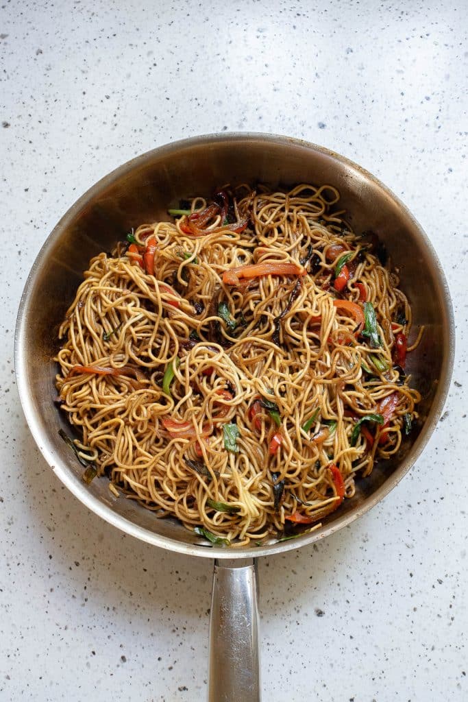 A stainless steel pan on a white countertop contains cooked noodles mixed with vegetables, including bell peppers and greens. The dish appears to be a stir-fry with a mix of ingredients providing a colorful and appetizing appearance.