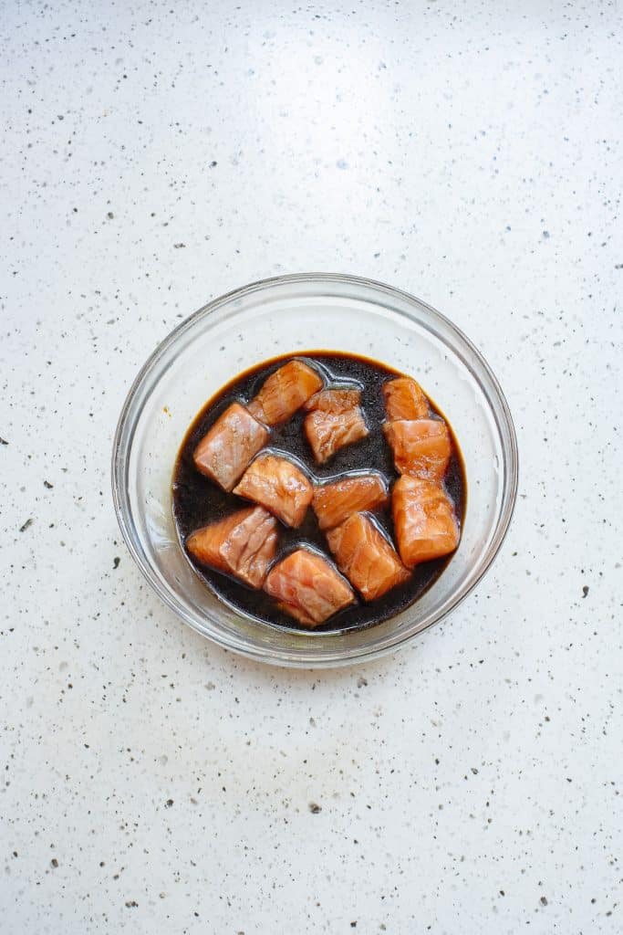 A clear glass bowl containing cubed pieces of raw salmon marinating in a dark liquid, likely soy sauce or similar, placed on a speckled white countertop.