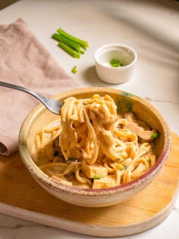 A bowl of creamy pasta garnished with green onions is set on a wooden board. A fork is lifting noodles from the bowl. A linen napkin and a small white ramekin with green onions are placed in the background on a marble countertop.