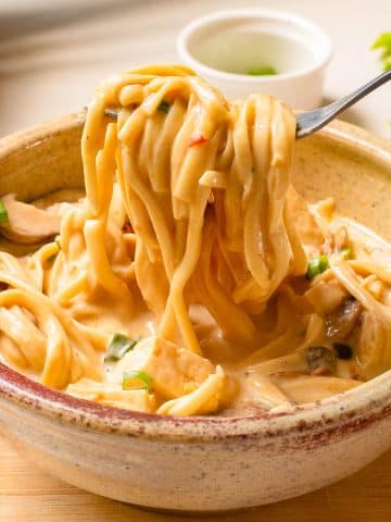 A bowl of creamy pasta with vegetables and mushrooms, held by a hand with a fork lifting a portion of noodles. The pasta is garnished with chopped herbs, placed on a wooden surface with a small white condiment bowl in the background.