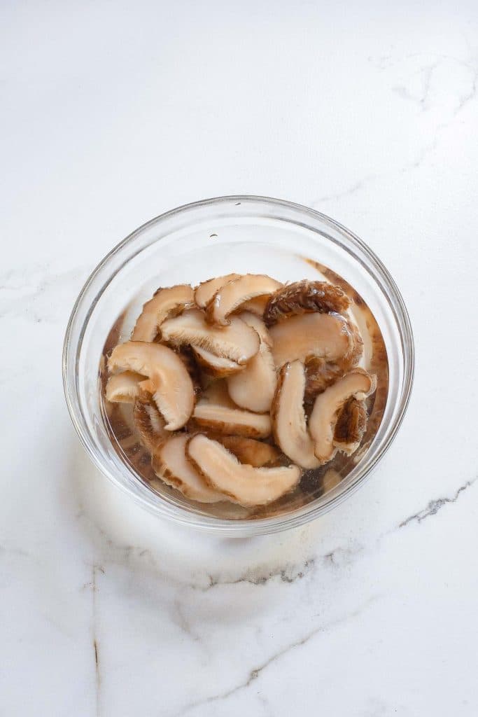 A clear glass bowl filled with sliced shiitake mushrooms soaking in water. The bowl is placed on a white marble countertop.
