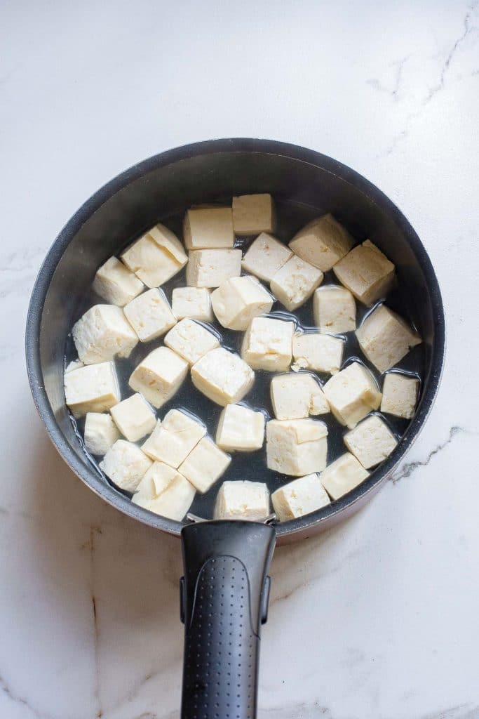 A black saucepan containing cubed pieces of tofu sits on a light-colored marble countertop. The tofu cubes are partially submerged in water.