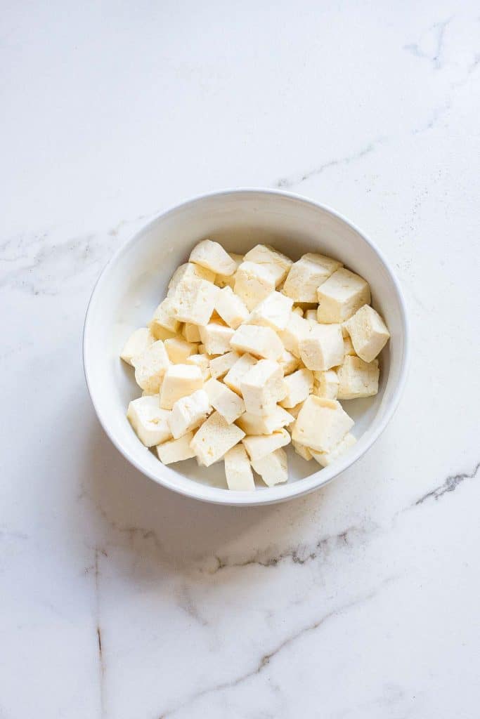 A white bowl filled with diced tofu cubes, placed on a marble countertop. The tofu pieces are uniform in size and pale in color, creating a simple and clean visual contrast with the white bowl and surface.