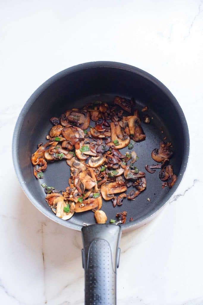 A round black pan with a black handle holds cooked sliced mushrooms and chopped green herbs. The pan is placed on a light-colored marble surface.