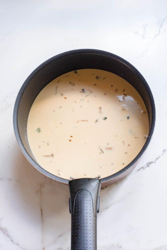 A black saucepan containing a creamy soup with visible pieces of mushrooms and herbs rests on a white marble surface.