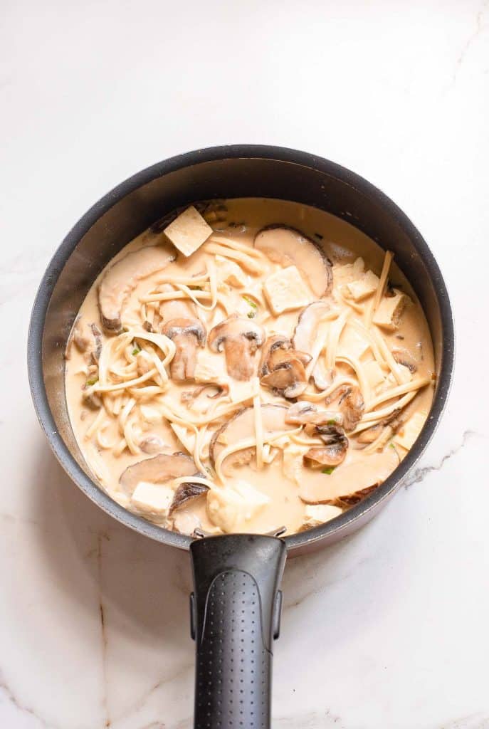 A black pan on a white marble surface contains a creamy mushroom soup with sliced mushrooms, tofu cubes, and noodles. The soup is light brown in color and appears to be rich and hearty.