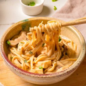 A bowl of creamy udon noodles is shown on a wooden surface. The noodles are topped with green onions and bits of tofu or chicken. A pair of wooden chopsticks is lifting a bundle of noodles from the bowl. A small ramekin with more green onions is in the background.