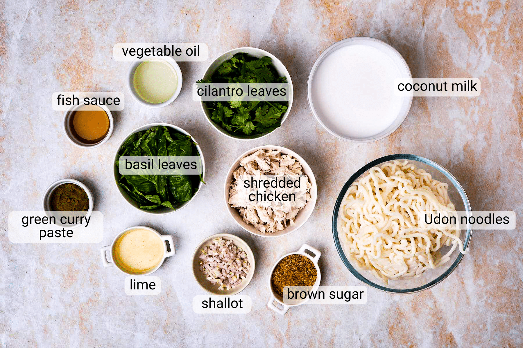 Top-down view of ingredients in bowls for a noodle dish, including udon noodles, shredded chicken, coconut milk, basil leaves, cilantro leaves, lime, green curry paste, fish sauce, vegetable oil, shallot, and brown sugar.