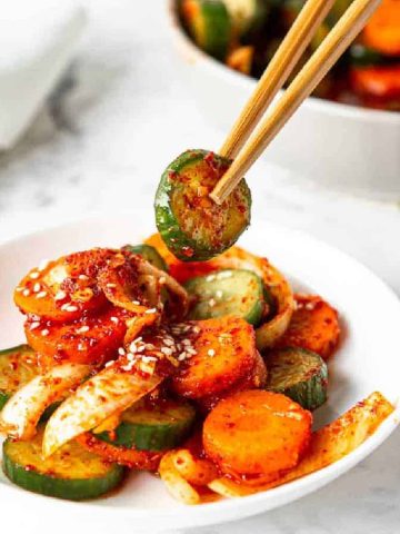 A bowl of spicy cucumber and carrot salad garnished with sesame seeds. Chopsticks hold a slice of cucumber above the bowl. There's a white napkin and another bowl of salad in the background on a marble surface.