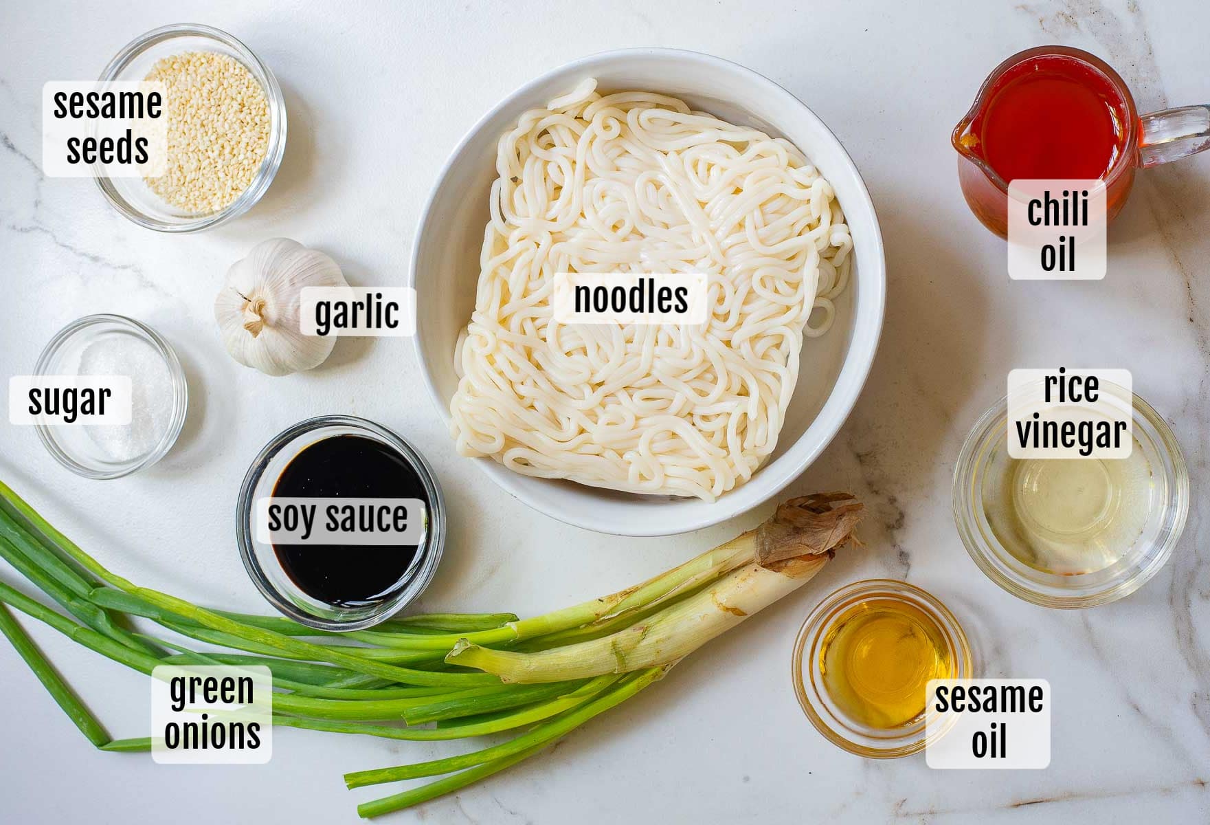 A top-down view of labeled ingredients for garlic chili oil noodles on a white surface: a bowl of noodles, garlic, green onions, sesame seeds, sugar, soy sauce, sesame oil, rice vinegar, and chili oil in bowls or small containers.