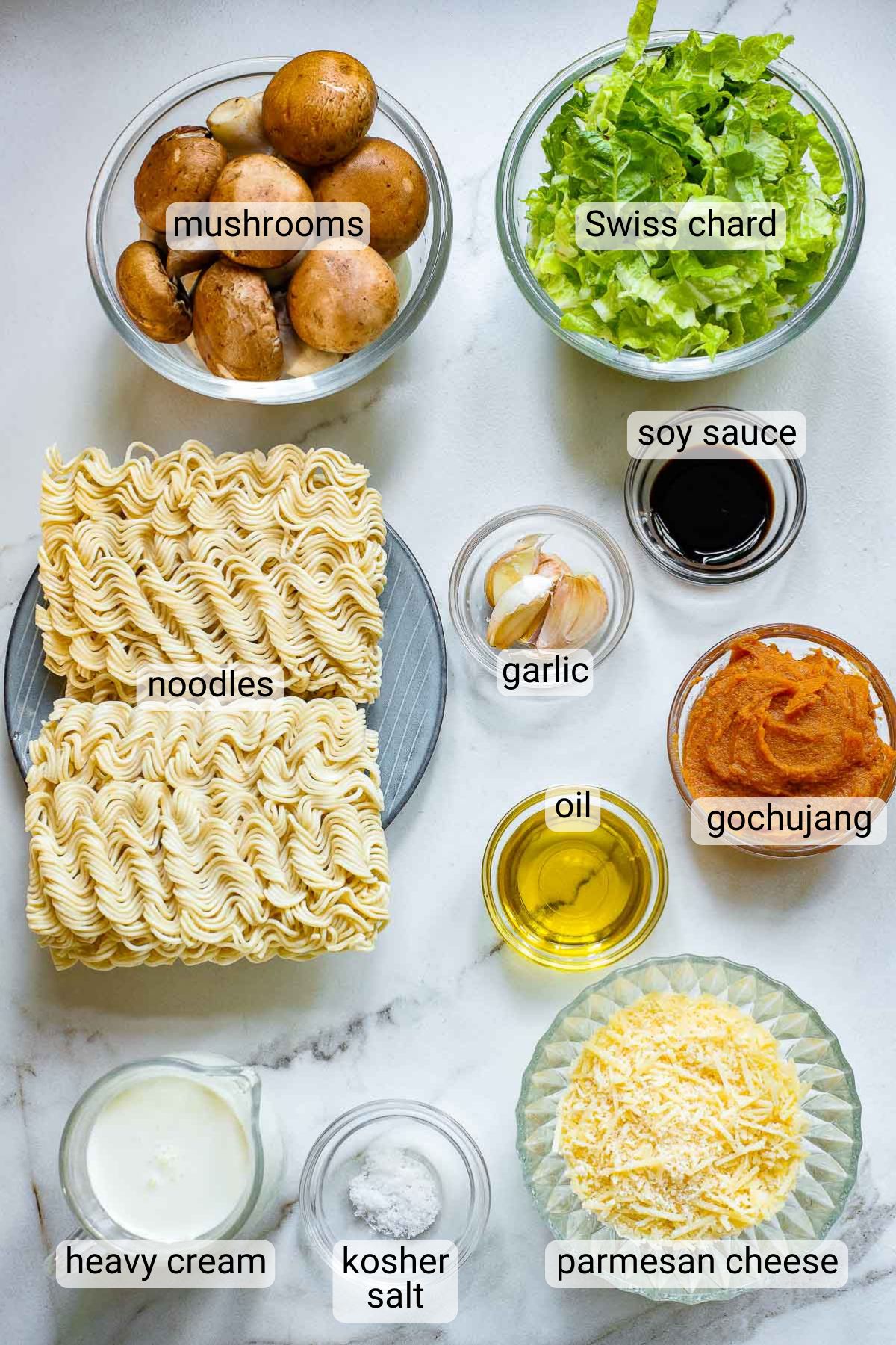Ingredients displayed on a table include labeled bowls of mushrooms, Swiss chard, soy sauce, garlic, gochujang, oil, heavy cream, kosher salt, parmesan cheese, and a plate with uncooked noodles.