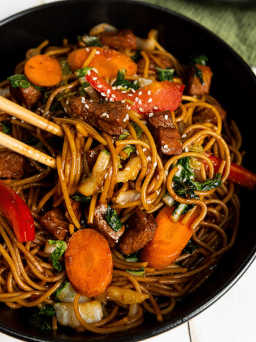 A hand holding chopsticks prepares to pick up a bite of stir-fried noodles mixed with sliced carrots, bell peppers, and greens from a black bowl. The dish appears colorful and appetizing, with bits of meat and sesame seeds sprinkled on top.