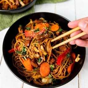A bowl of yakisoba noodles with vegetables and pieces of meat, garnished with sesame seeds. A hand with chopsticks is about to pick up some noodles. There are sliced carrots, red bell peppers, and greens mixed with the noodles. Another bowl is in the background.