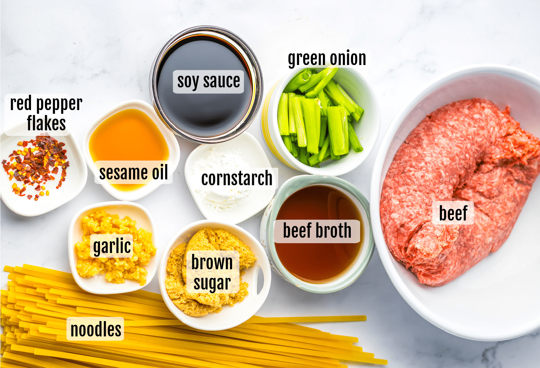 Ingredients for a beef noodle dish arranged on a white surface, including ground beef, uncooked noodles, green onion, soy sauce, beef broth, garlic, brown sugar, sesame oil, cornstarch, and red pepper flakes, each labeled.