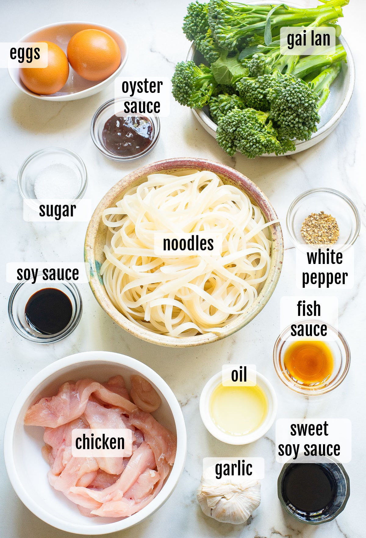 Ingredients for a noodle dish arranged on a marble surface: eggs, gai lan (Chinese broccoli), bowls of oyster sauce, sugar, soy sauce, noodles, white pepper, fish sauce, sweet soy sauce, oil, sliced chicken, and garlic.