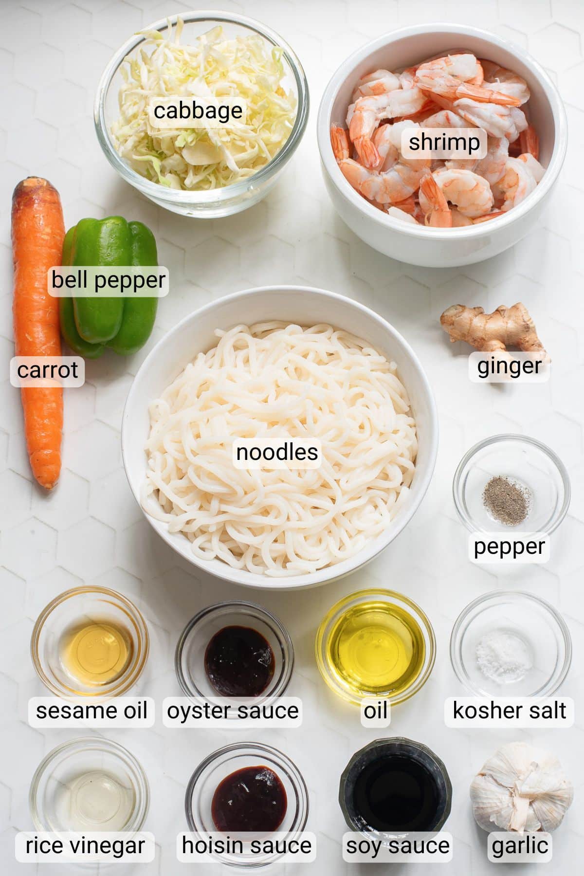 Overhead view of ingredients for a noodle dish, including sliced cabbage, shrimp, ginger, bell pepper, carrot, noodles, and small bowls of sesame oil, oyster sauce, oil, hoisin sauce, soy sauce, rice vinegar, garlic, pepper, and kosher salt.
