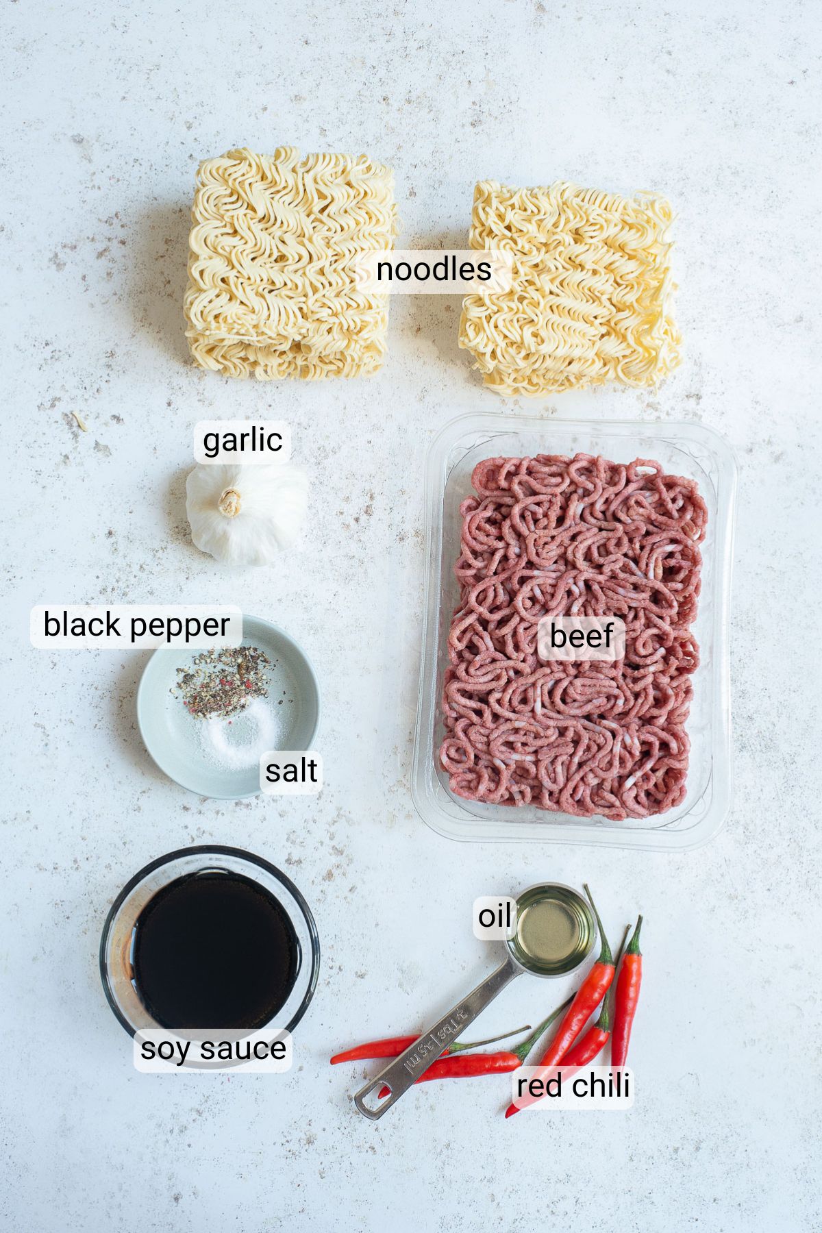 Ingredients laid out on a surface: two blocks of noodles, a head of garlic, a dish of black pepper and salt, a container of ground beef, a bowl of soy sauce, a tablespoon of oil, and several red chilies.