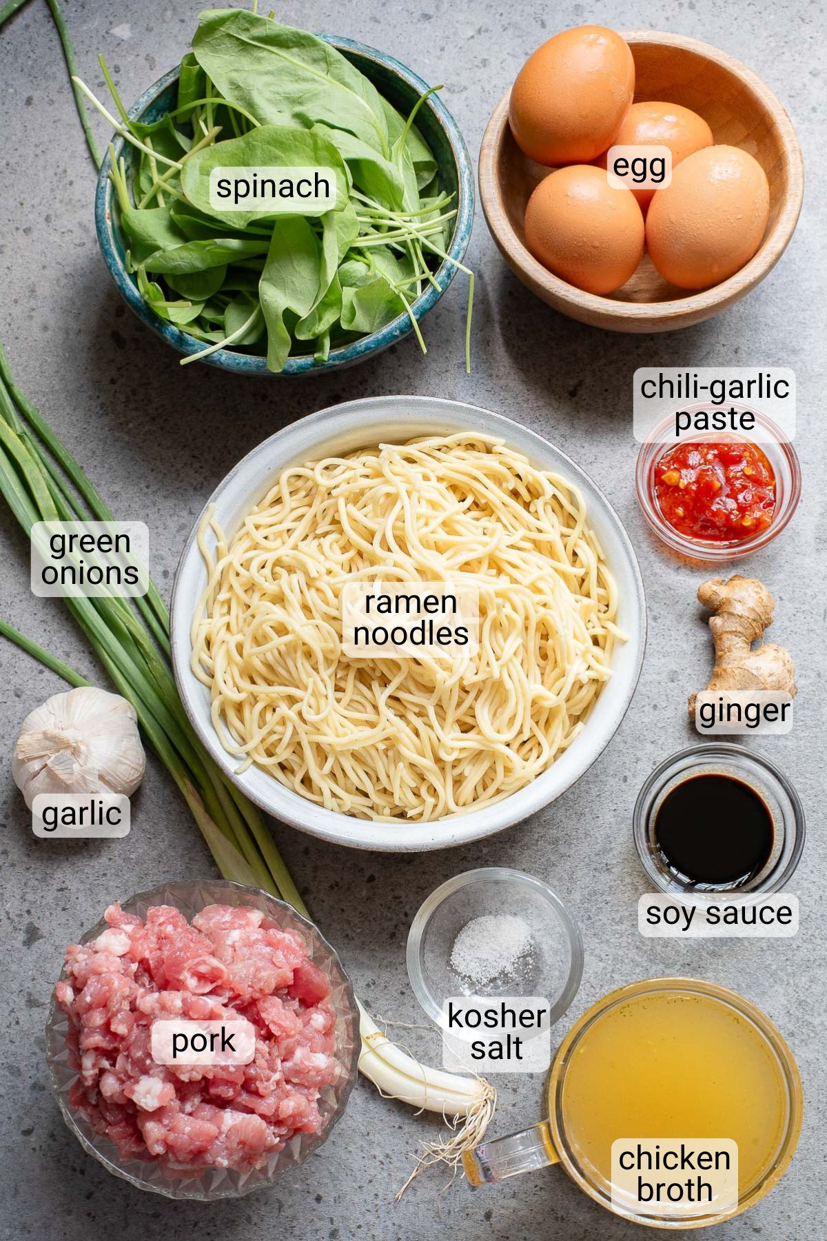 A flat lay of ingredients for ramen: spinach, eggs, green onions, ramen noodles, chili-garlic paste, garlic, pork, ginger, soy sauce, kosher salt, and chicken broth on a gray surface.