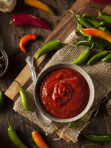 A bowl of red hot sauce sits on a rustic wooden surface, embodying the spicy food trend. It's surrounded by colorful chili peppers, a spoon, and burlap cloth. Garlic cloves and peppercorns are scattered around, enhancing the fiery ambiance.