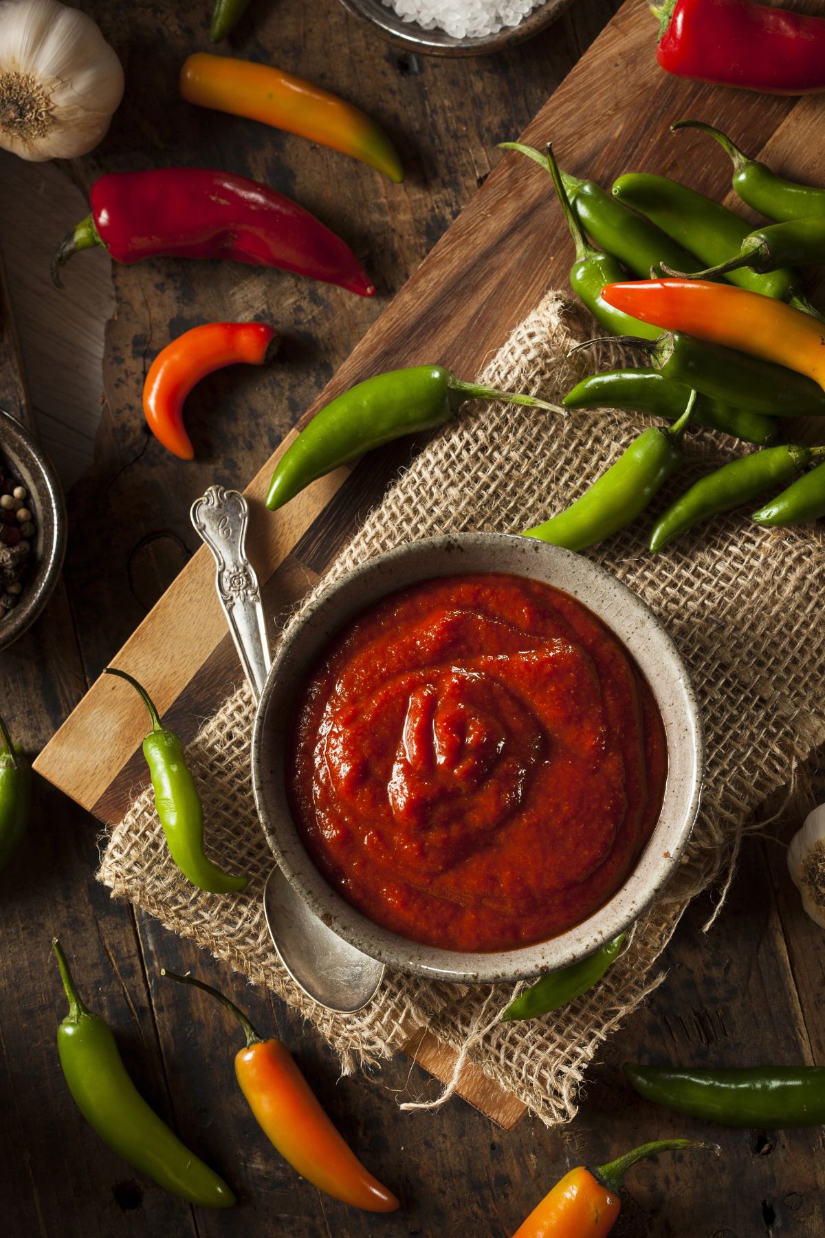 A bowl of red hot sauce sits on a rustic wooden surface, embodying the spicy food trend. It's surrounded by colorful chili peppers, a spoon, and burlap cloth. Garlic cloves and peppercorns are scattered around, enhancing the fiery ambiance.