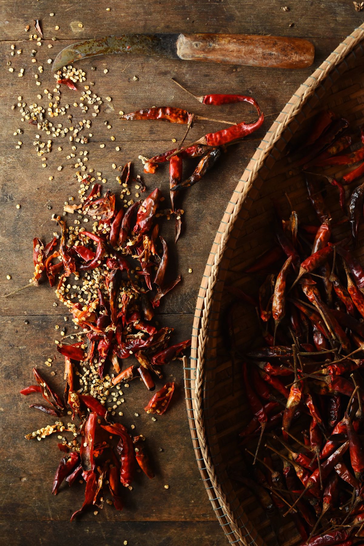 Dried red chilies and scattered seeds are on a wooden surface next to a woven basket filled with more chilies, capturing the essence of the spicy food trend. A rustic knife with a wooden handle is placed nearby.
