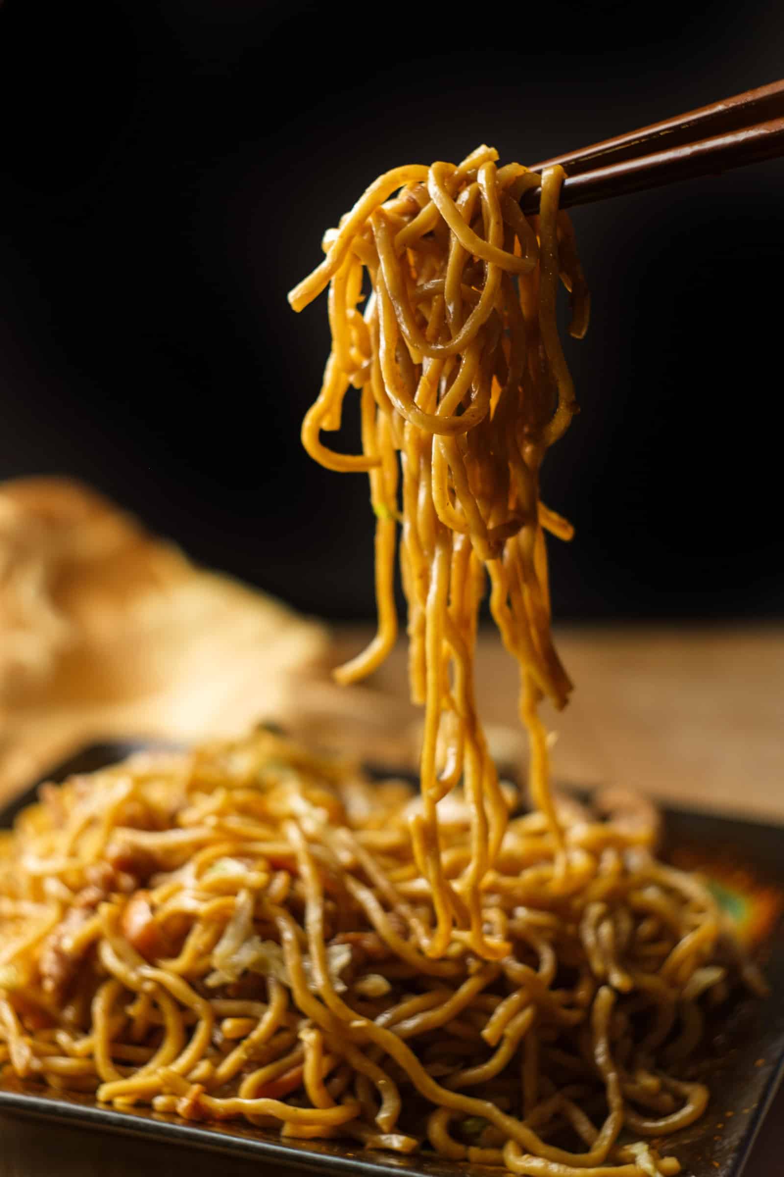Close-up of chopsticks lifting a portion of Asian noodles from a plate against a dark background. The golden noodles appear seasoned, with blurred portions of the dish visible below, offering an inviting glimpse into this culinary delight.