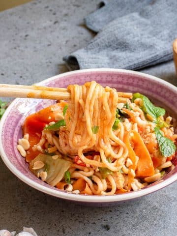 A bowl of spicy noodles with vegetables and herbs, garnished with crushed peanuts. A pair of chopsticks holds a bundle of noodles above the bowl. The dish is served on a speckled gray surface, with a gray cloth in the background.