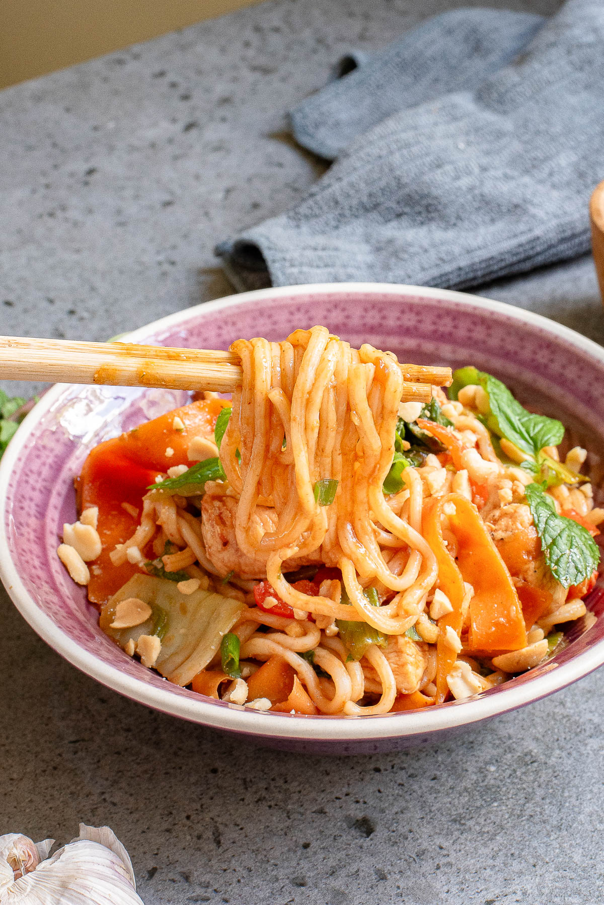 A bowl of spicy noodles with vegetables and herbs, garnished with crushed peanuts. A pair of chopsticks holds a bundle of noodles above the bowl. The dish is served on a speckled gray surface, with a gray cloth in the background.