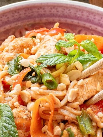 A bowl of noodle stir-fry topped with vegetables, chicken, crushed peanuts, and fresh mint leaves. Chopsticks rest on the side of the bowl, which is placed on a textured surface with a small container in the background.