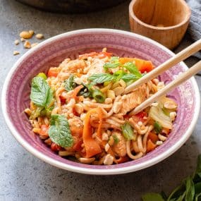 A bowl of colorful stir-fried noodles with vegetables, tofu, peanuts, and fresh basil leaves. Wooden chopsticks rest on the rim, and a small wooden bowl is nearby. The dish is served in a purple and white bowl on a gray surface.