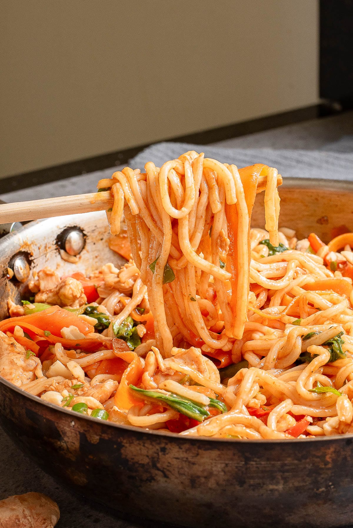 A fork lifting a portion of stir-fried noodles from a pan. The noodles are mixed with colorful vegetables like carrots and bell peppers, and small pieces of meat, seasoned with a savory sauce.