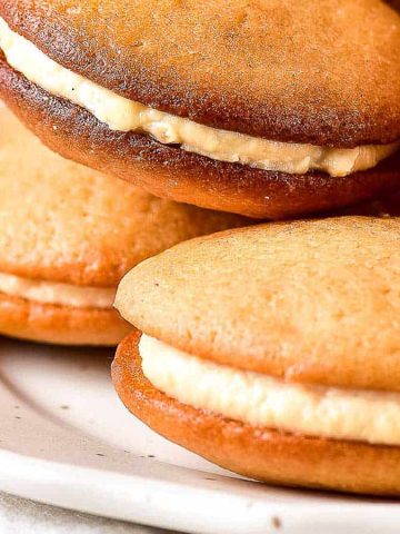 A plate of freshly baked whoopie pies with golden brown tops and creamy filling. The whoopie pies are stacked and layered, showcasing their soft and inviting texture.