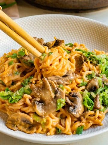 A bowl of creamy mushroom ramen topped with green onions on a textured plate. Chopsticks are lifting noodles, highlighting the delicious texture and rich color of the dish.