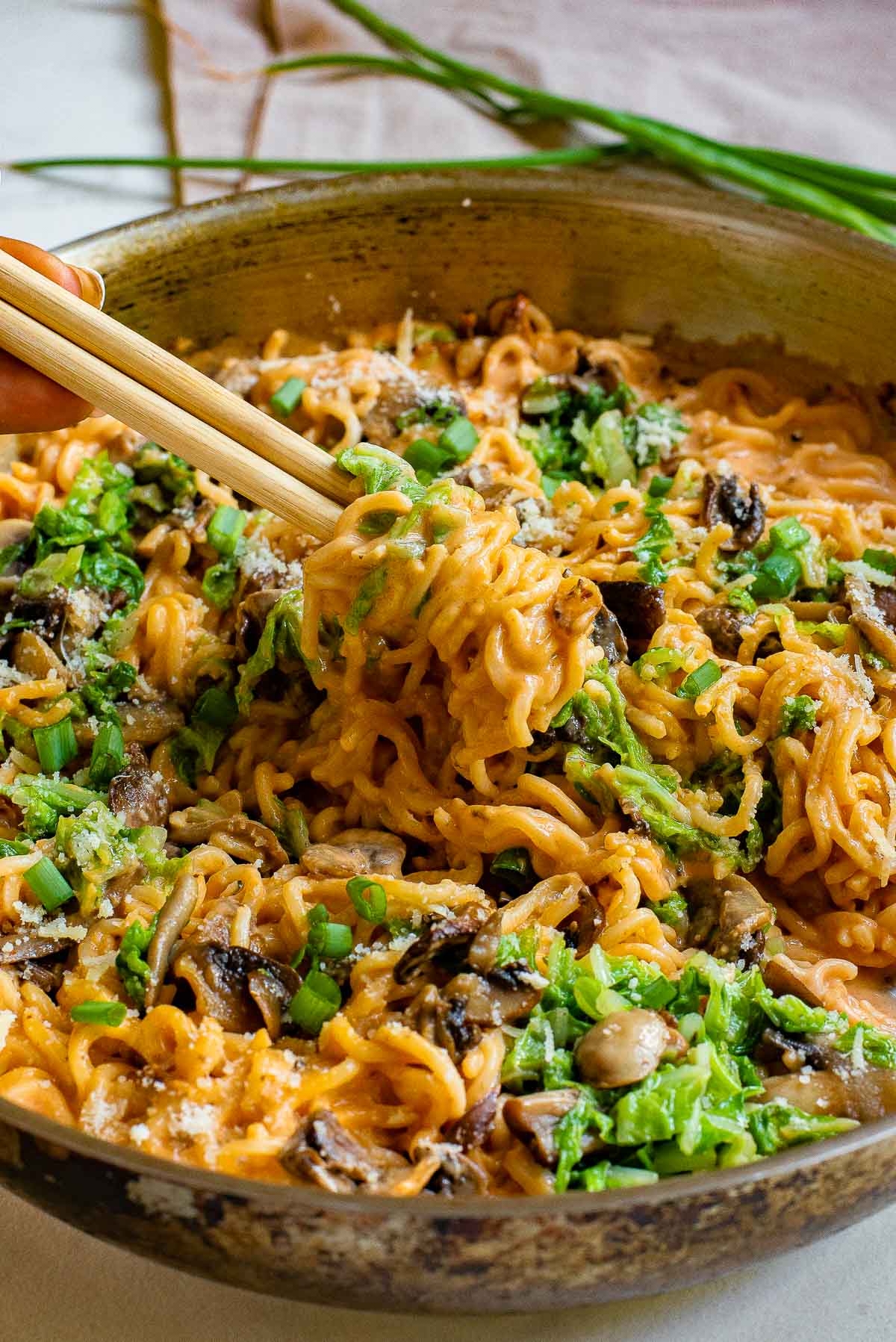 A bowl of creamy noodles mixed with mushrooms and chopped green vegetables. Chopsticks are lifting a portion of the noodles. The dish is garnished with a sprinkle of grated cheese. Green onions are placed in the background.