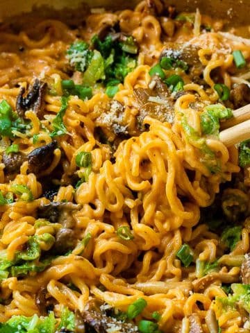 A bowl of creamy ramen noodles mixed with mushrooms and topped with green onions and herbs. Chopsticks are lifting a portion of the noodles. The dish is served in a shallow pan.