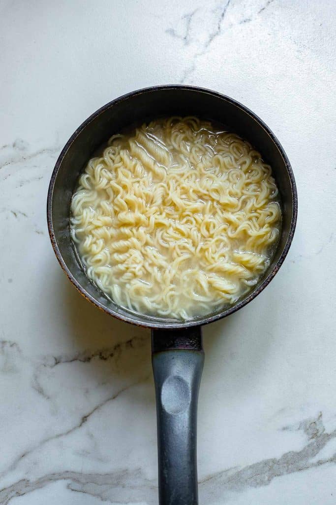 A small black saucepan filled with cooked ramen noodles in a light broth sits on a white marble surface. The ramen is arranged in a wavy pattern, and the handle of the saucepan is visible in the foreground.